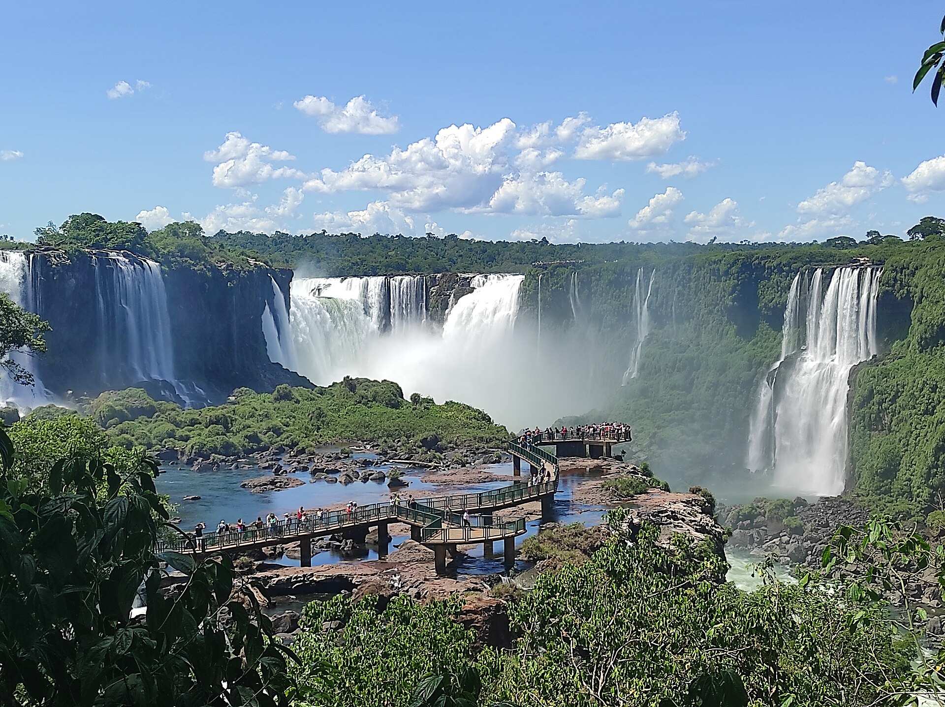 Quedas-d'água das Cataratas do Iguaçu vistas do lado brasileiro, com passarela e visitantes entre a mata e o céu azul