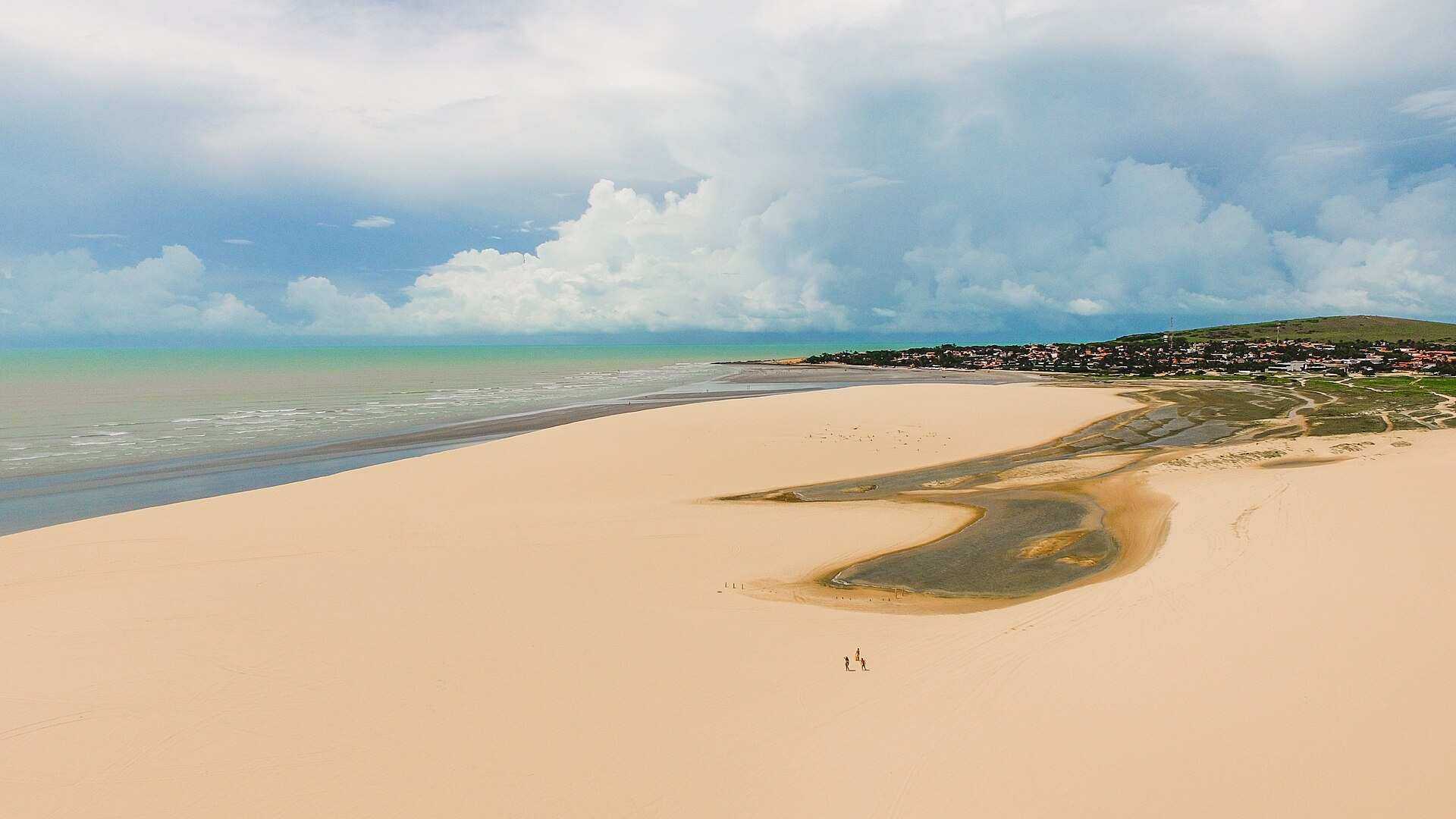Vista aérea de uma grande duna de areia clara com o mar à esquerda e a vila ao fundo