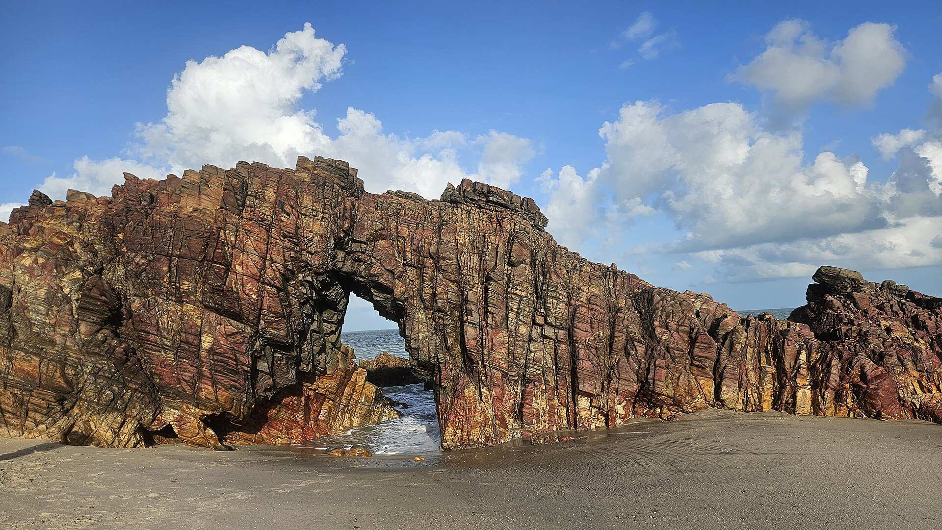 A Pedra Furada, arco de rocha avermelhada na praia, com o mar visível pela abertura