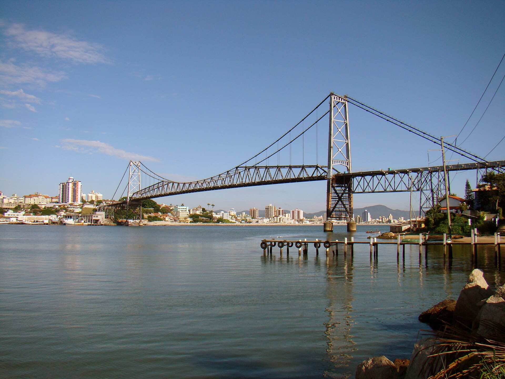 Ponte pênsil Hercílio Luz vista da margem, com a baía e a cidade ao fundo sob céu azul